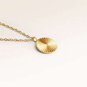 Close-up of minimalist round pendant in gold with ribbed sunburst surface and space for engraving on back